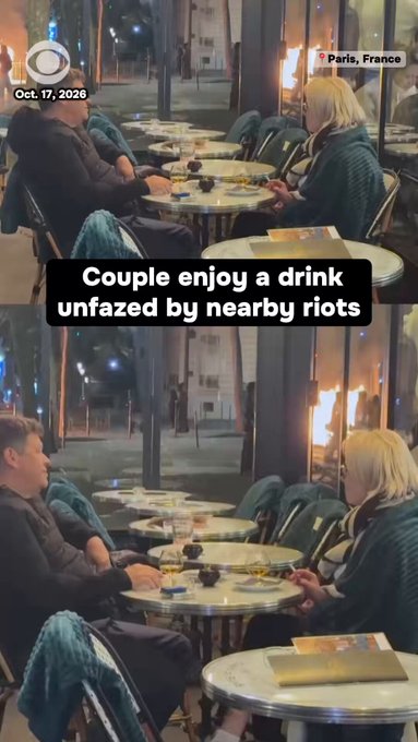 Pareja disfrutando de una bebida en la terraza de un café cerca del Arco de Triunfo mientras arden las calles de París.